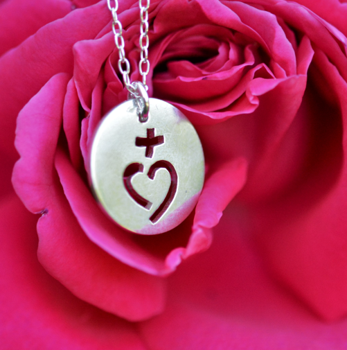 un pendentif en argent ajouré d'un coeur sacré est posé devant une rose.