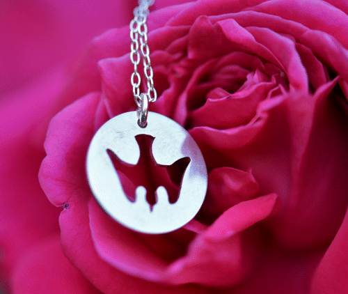 un pendentif en argent ajouré d'une colombe est posé devant une rose.