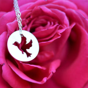 un pendentif en argent ajouré d'une colombe est posé devant une rose.