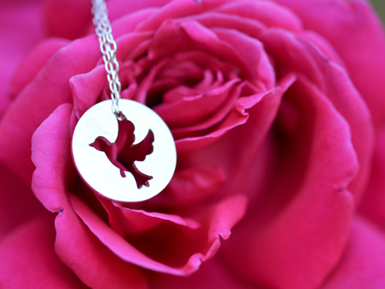 un pendentif en argent ajouré d'une colombe est posé devant une rose.
