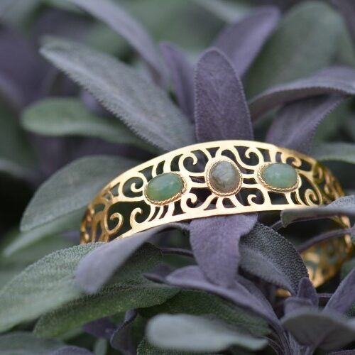 Manchette Couleurs de sauge dorée ajourée posée dans une plante, avec des pierres d'aventurine et de labradorite.